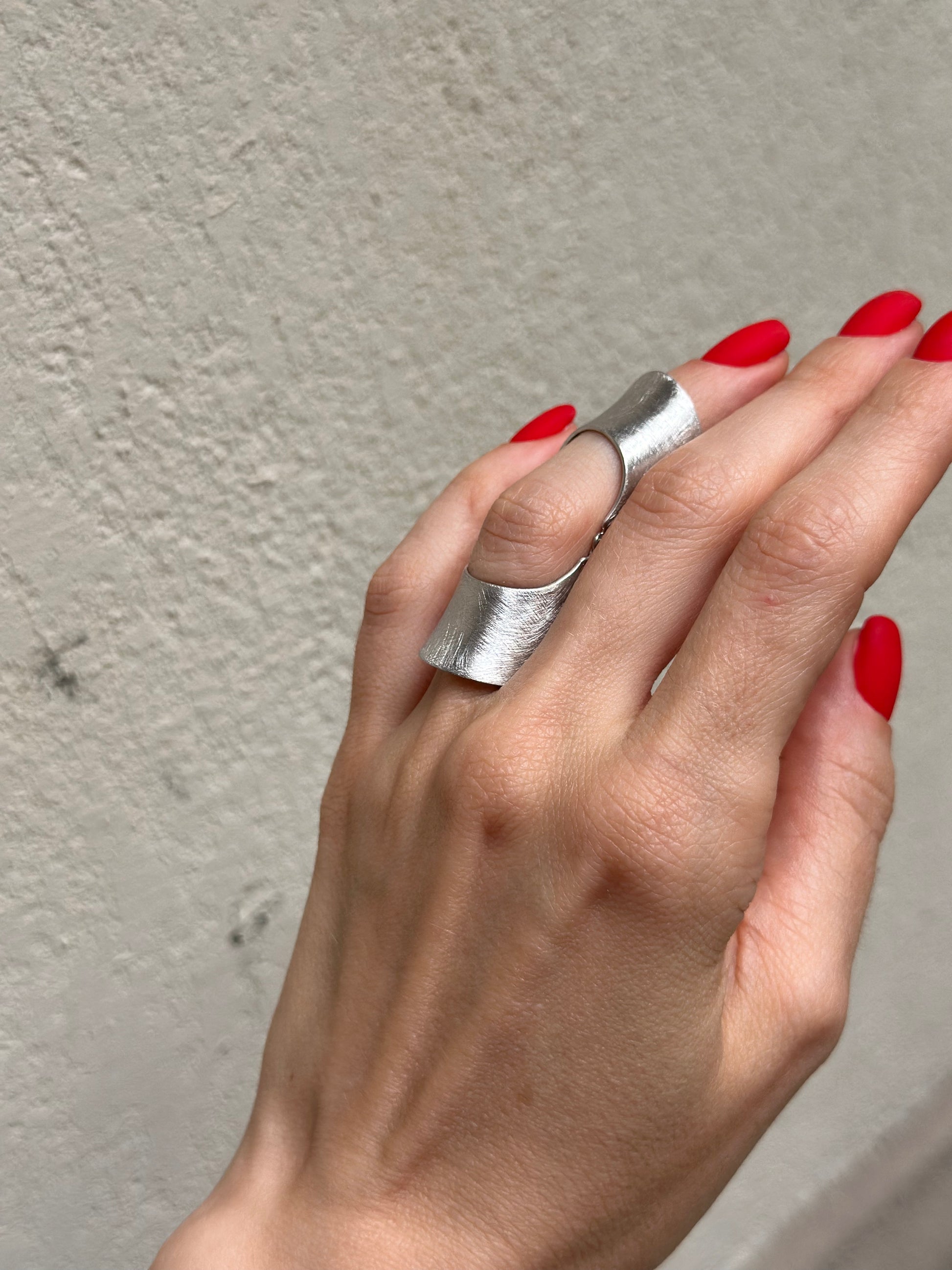 Hand wearing a silver ring with red nail polish against a textured wall.
