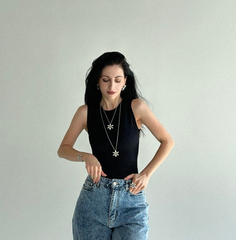 Woman wearing a black sleeveless top and blue jeans with floral jewelry, long floral necklace, ring, earrings and bracelet, against a plain background