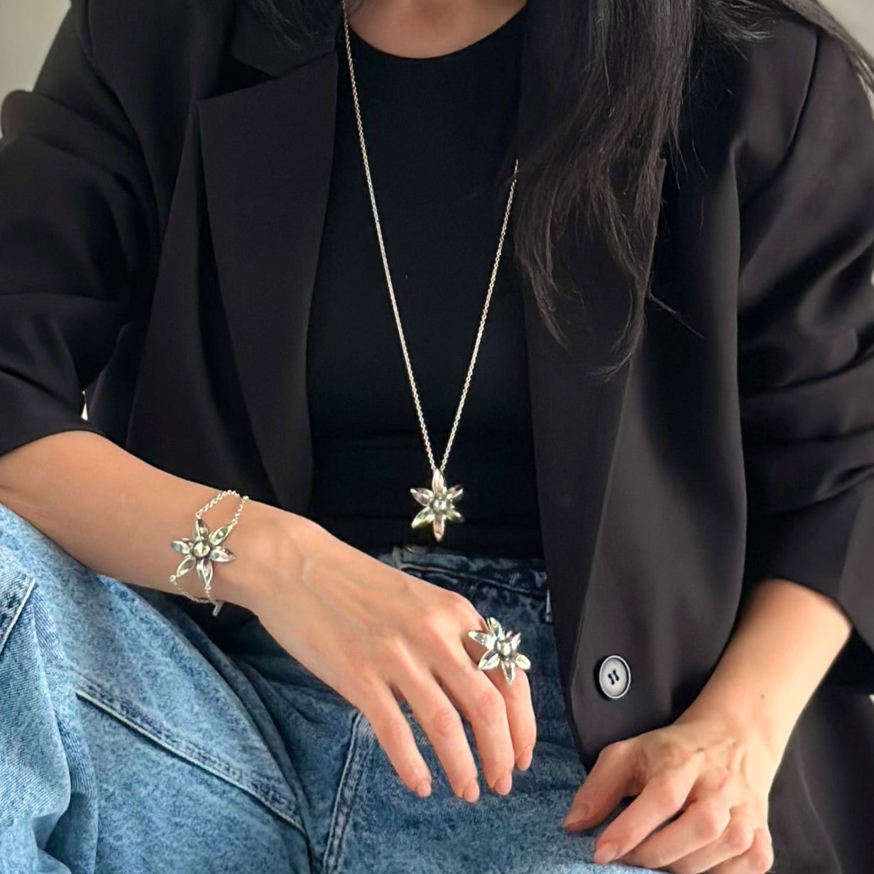 Woman sitting on a staircase wearing a black coat, black top, and blue jeans. with flower jewelry ring flower, bracelet flower and necklace flower